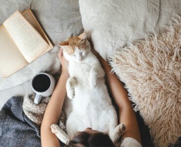cat sleeping in woman's lap with open book and coffee mug